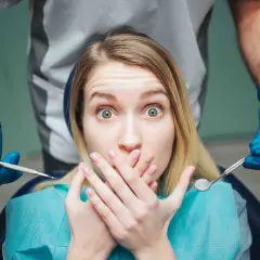 Occhi sbarrati e mani sulla bocca: la paura del dentista è palpabile mentre il dentista si avvicina con gli strumenti.