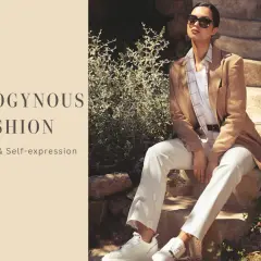 Modello androgino con blazer cammello, pantaloni bianchi e sneakers, su gradini di pietra.