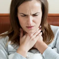 Donna con mani sul collo, espressione sofferente, forse per difficoltà a deglutire o ansia.