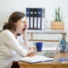 Donna al lavoro, con sbadigli continui, forse per l'ansia o la stanchezza. Sul tavolo, laptop, tazza blu e quaderni.