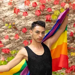 Un giovane con la bandiera arcobaleno, simbolo del **queer significato** di libertà e identità, si staglia contro un muro fiorito.