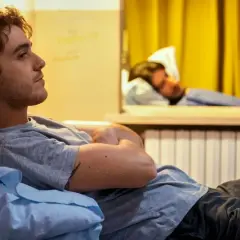 Un giovane uomo pensieroso, forse un genio incompreso o in preda alla follia, riposa su un letto in una stanza con tende gialle.