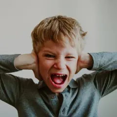 Bambino con le mani sulle orecchie, urla con espressione di rabbia, possibile manifestazione di disturbi del comportamento.