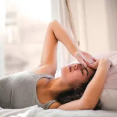 Ragazza sdraiata a letto, con la mano sulla fronte, sembra che non riesca a svegliarsi la mattina. Luce soffusa dalla finestra.