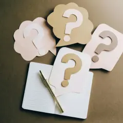 Penne dorata su quaderno aperto, circondata da fumetti con punti interrogativi, simbolo di pensieri automatici negativi e dubbi.