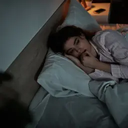 Una donna in pigiama, a letto, si morde le unghie. Sono stanca ma non riesco a dormire, il buio della stanza accentua l'ansia.