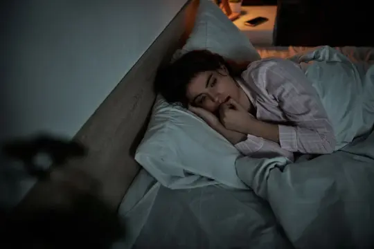 Una donna in pigiama, a letto, si morde le unghie. Sono stanca ma non riesco a dormire, il buio della stanza accentua l'ansia.