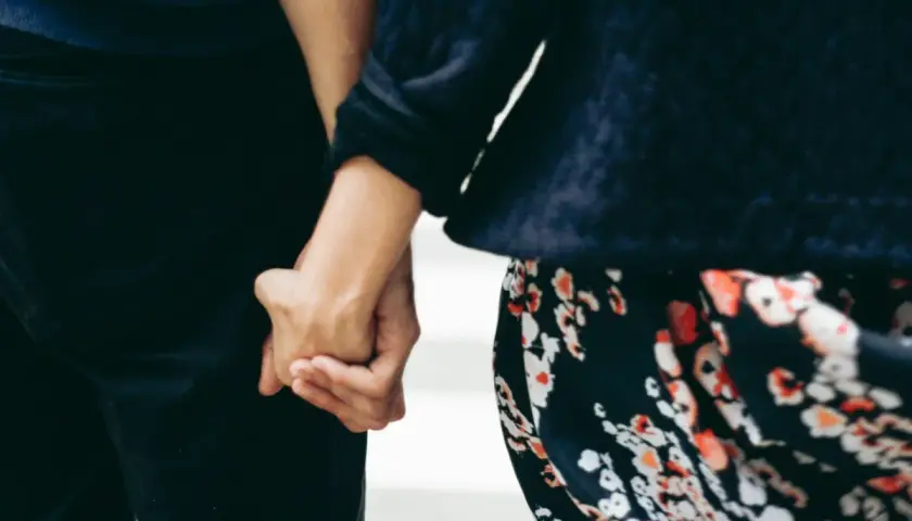 Mani intrecciate, un gesto che parla di affinità di coppia e complicità. La donna indossa un abito floreale, l'uomo pantaloni scuri.
