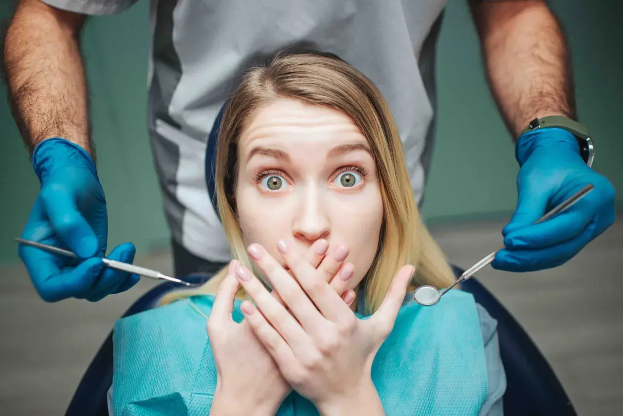 Occhi sbarrati e mani sulla bocca: la paura del dentista è palpabile mentre il dentista si avvicina con gli strumenti.