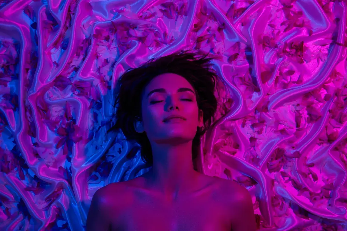 Donna con occhi chiusi, immersa in un letto di seta e petali, illuminata da luci blu e rosa. Un'immagine che evoca il piacere e il mistero del perché godiamo quando veniamo.