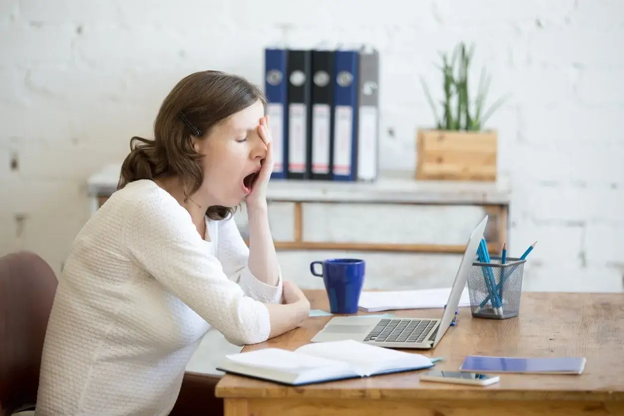 Donna al lavoro, con sbadigli continui, forse per l'ansia o la stanchezza. Sul tavolo, laptop, tazza blu e quaderni.