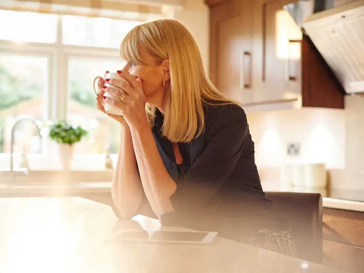 Donna bionda con tazza di caffè riflette sulla sua crisi di mezza età, con luce solare che filtra in cucina.
