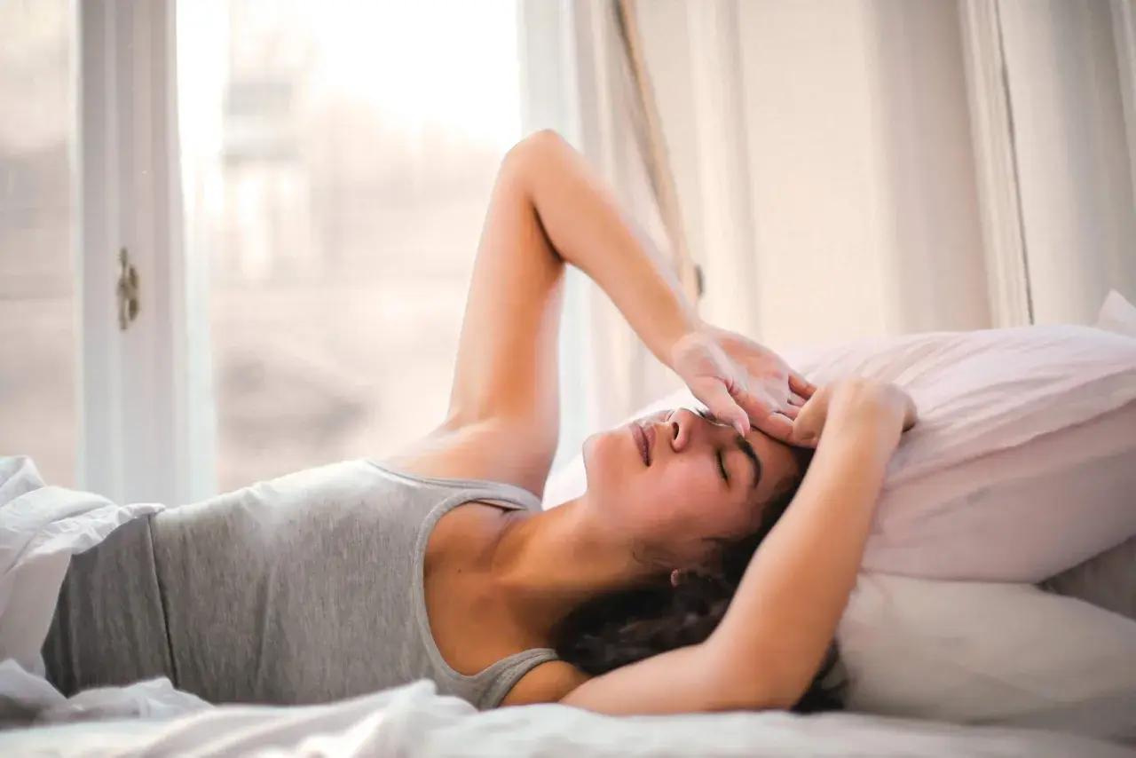 Ragazza sdraiata a letto, con la mano sulla fronte, sembra che non riesca a svegliarsi la mattina. Luce soffusa dalla finestra.