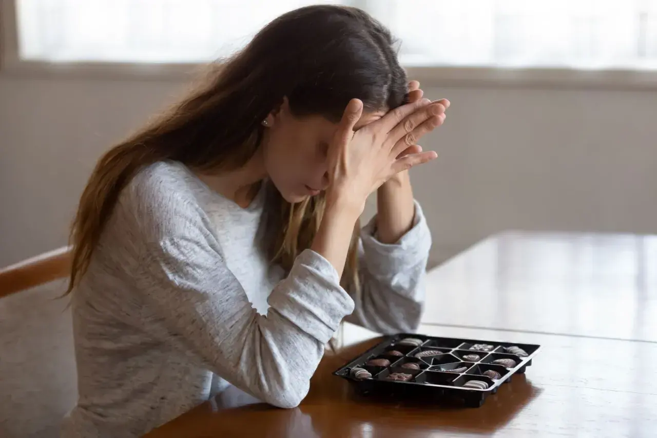 Donna con le mani sul viso, davanti a una scatola di cioccolatini, forse cercando un antidepressivo naturale più potente.
