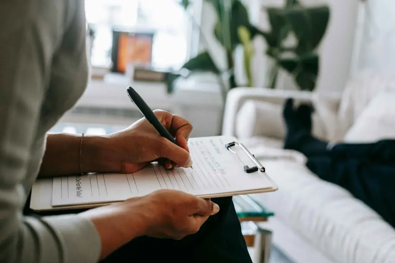 Mani che scrivono su un blocco note, mentre una persona è sdraiata su un divano. Un momento di riflessione per capire se per l'ansia meglio psicologo o psichiatra.
