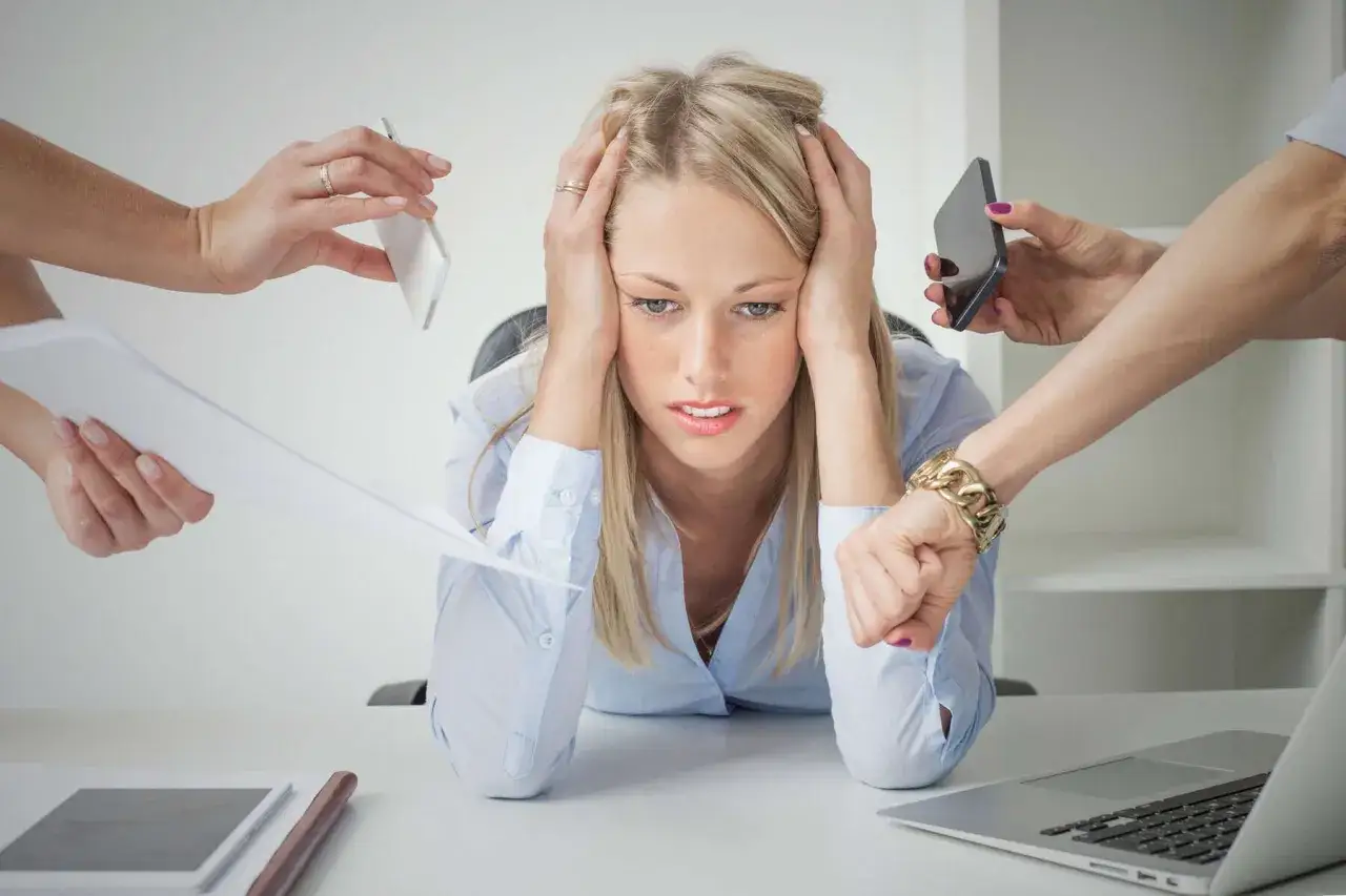 Donna stressata con mani tra i capelli, circondata da mani che le porgono telefoni e documenti. Sintomo di dipendenza da lavoro.