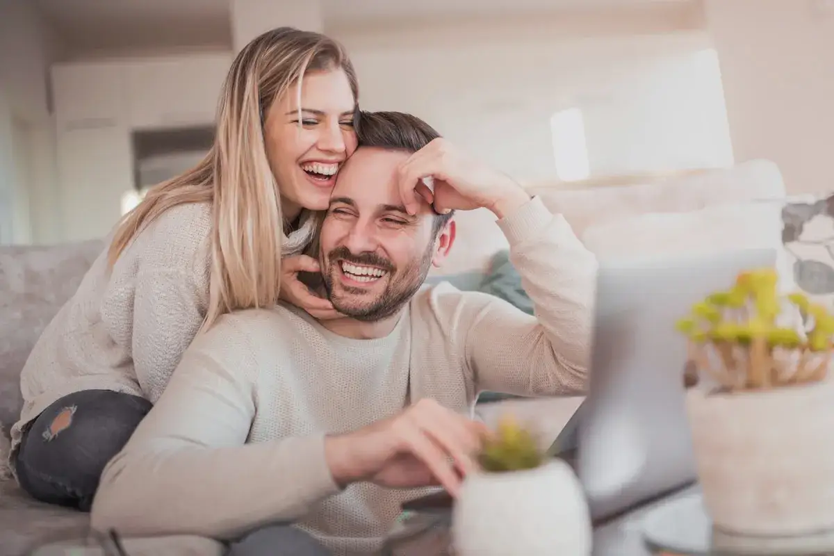 Coppia felice che ride sul divano, lei lo abbraccia da dietro. Mio marito vuole fare l'amore tutti i giorni, e momenti cos&igrave; lo dimostrano.