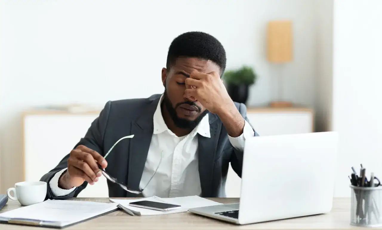 Uomo d'affari con la mano sulla fronte, occhiali in mano, laptop e tazza di caffè sulla scrivania. Sembra soffrire di ansia ad alto funzionamento.
