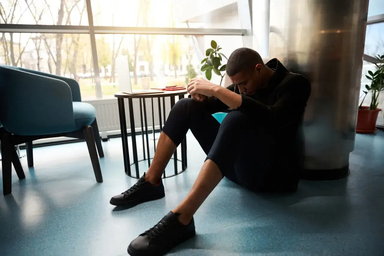 Un giovane uomo seduto a terra, con la testa china, sembra riflettere. Vicino a lui, una sedia blu e un tavolino con un laptop e quaderni. Piccoli segnali di ripresa dalla depressione.