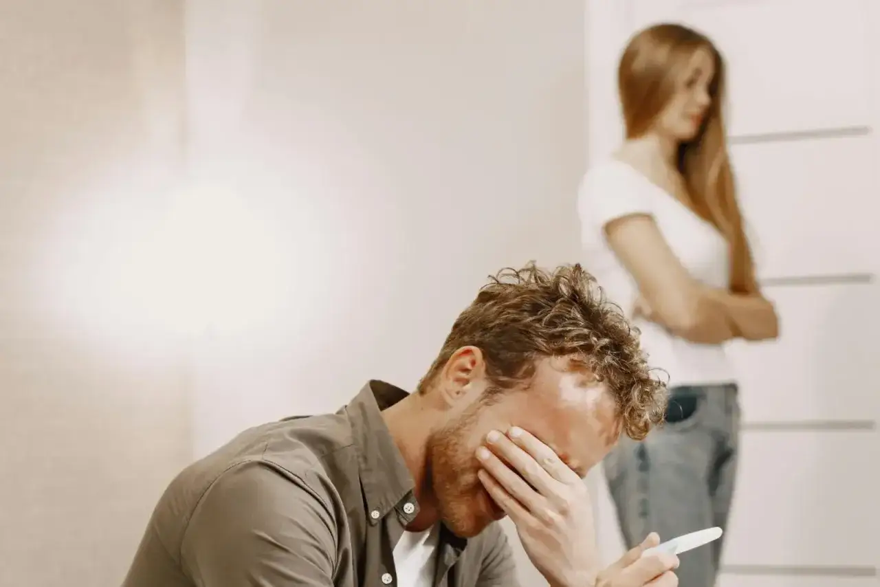 Uomo disperato con test di gravidanza in mano, donna con braccia conserte sullo sfondo. Forse si pentir&agrave; di credere di non amare pi&ugrave;.