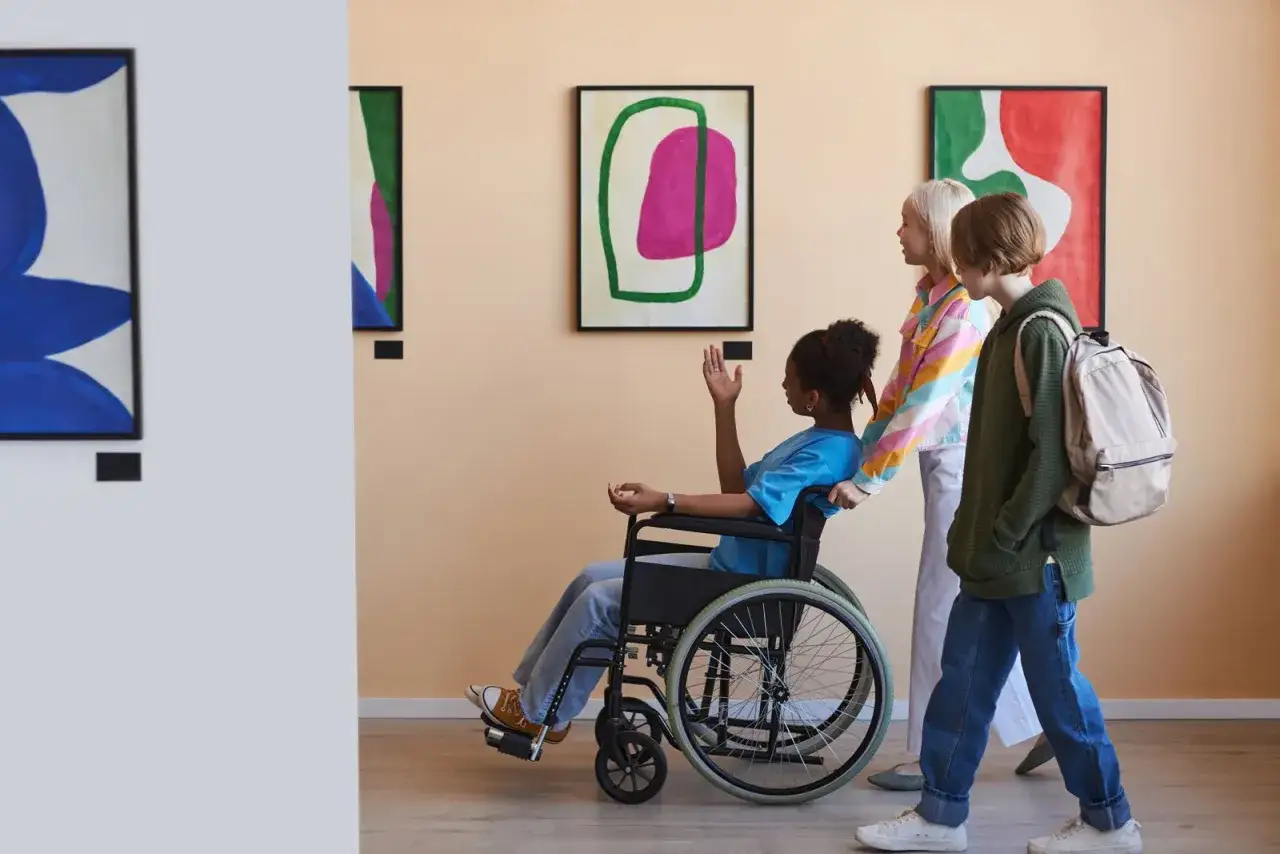 Bambini in galleria d'arte, tra cui una in sedia a rotelle, ammirano quadri astratti. Un'esplosione di genio e follia colorata.