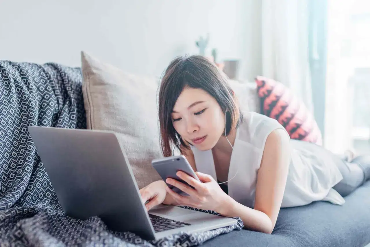Donna con mania del controllo, sdraiata sul divano, usa laptop e smartphone contemporaneamente.